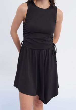 Frau in einem ärmellosen schwarzen Kleid mit seitlichen Raffungen und asymmetrischem Saum, die vor einem schlichten hellen Hintergrund steht.