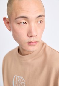 Beženi dukserica kratkih rukava s okruglim izrezom i svijetloplavim grafičkim detaljem. Glatka tekstura, nosi je osoba s obrijanom glavom.