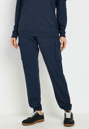 Marineblauw ribbed top en bijpassende cargo-broek met zijzakken en elastische manchetten; gecombineerd met zwarte sneakers met witte veters en een bruine zool.