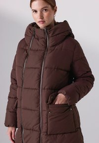 Pruun pehmendatud parka kõrge krae ja kapuutsiga, esiküljel tõmblukk ja kaks klapiga taskut. Tekstuuriga tikand.