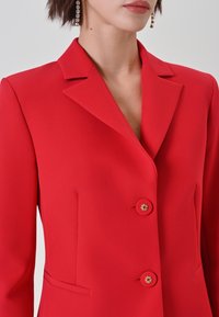 Blazer rosso con colletto a lancia, due chiusure con bottoni dorati e tasche laterali; realizzato in tessuto liscio con una vestibilità sartoriale.