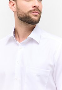 Een wit overhemd met een gestructureerde stof, knoopsluiting aan de voorkant en een enkele borstzak. De kraag heeft een geruit accent in blauw en rood.