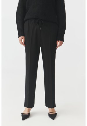 Black tailored trousers with a drawstring waist, straight leg design, and sleek fabric. Paired with pointed black flats and a textured black sweater.