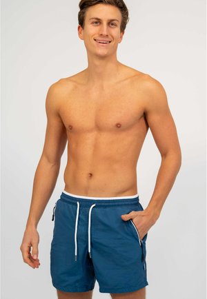 Jonge man glimlacht, bovenlichaam ontbloot, draagt blauwe zwemshorts met trekkoord en ritssluitingen in de zakken, staand tegen een effen lichte achtergrond.
