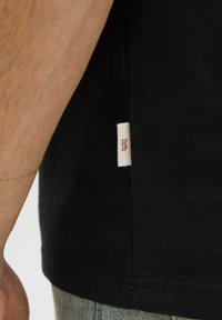 Sort bomulds t-shirt, der viser en sidevisning med et lille hvidt mærke med røde bølgede linjer nær kanten. Teksturen er glat.
