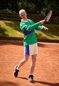 Teniszjátékos élénk zöld, hosszú ujjú pólóban, lila csíkkal és világos rövidnadrágban, ütőt tart a salakpályán. Fekete sportcipőt visel.