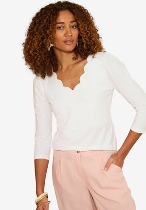 Vrouw met krullend haar die een wit topje met een geschulpte halslijn en lichtroze broek draagt, met één hand in de zak en armbanden om de pols.