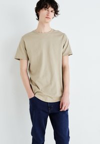 Beżowy bawełniany t-shirt z krótkimi rękawami i okrągłym dekoltem, zestawiony z ciemnoniebieskimi dżinsami. Prosty, luźny krój bez widocznych logo.