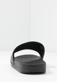Sandale noire à glissière avec une tige lisse et texturée, conception ouverte à l'avant, et une légère semelle compensée. Présente un style minimaliste et une sangle ajustable.