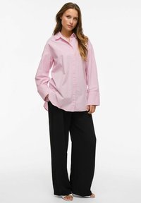 Šviesiai rožinė mygtukų marškiniai ilgomis rankovėmis, derinami su juodomis plačiomis kelnėmis, prieš baltą vientisą foną.