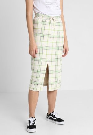Femme portant un T-shirt blanc et une jupe crème mi-genoux à carreaux verts et bleus, fente à l'avant, et des baskets noires avec des lacets blancs.