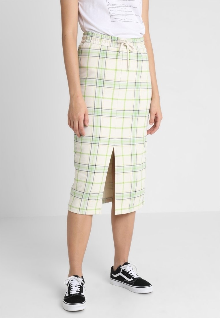 Femme portant un T-shirt blanc et une jupe crème mi-genoux à carreaux verts et bleus, fente à l'avant, et des baskets noires avec des lacets blancs.