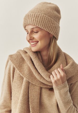 Femme souriante portant un bonnet en tricot beige, une écharpe assortie drapée autour de ses épaules et un pull beige, sur un fond clair uni.