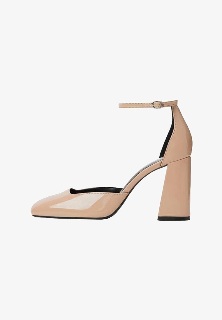 Chaussure à talon haut en cuir verni beige avec bout pointu fermé et bride de cheville avec petite boucle sur fond blanc.