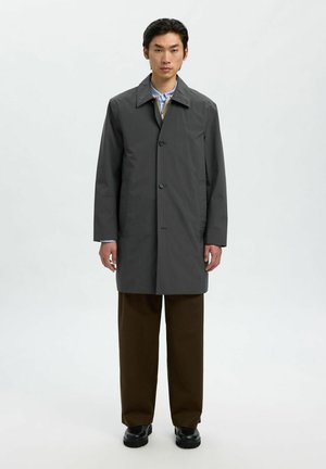 Man die recht vooruit kijkt en een donkergrijze button-up jas, bruine broek, zwarte schoenen en een gelaagde blouse met kraag draagt tegen een effen achtergrond.