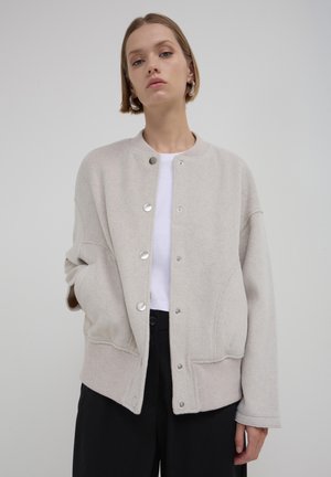 Veste en mélange de laine beige avec un ourlet côtelé, fermeture à pression et coutures courbes. Caractéristiques : coupe décontractée et épaules tombantes.