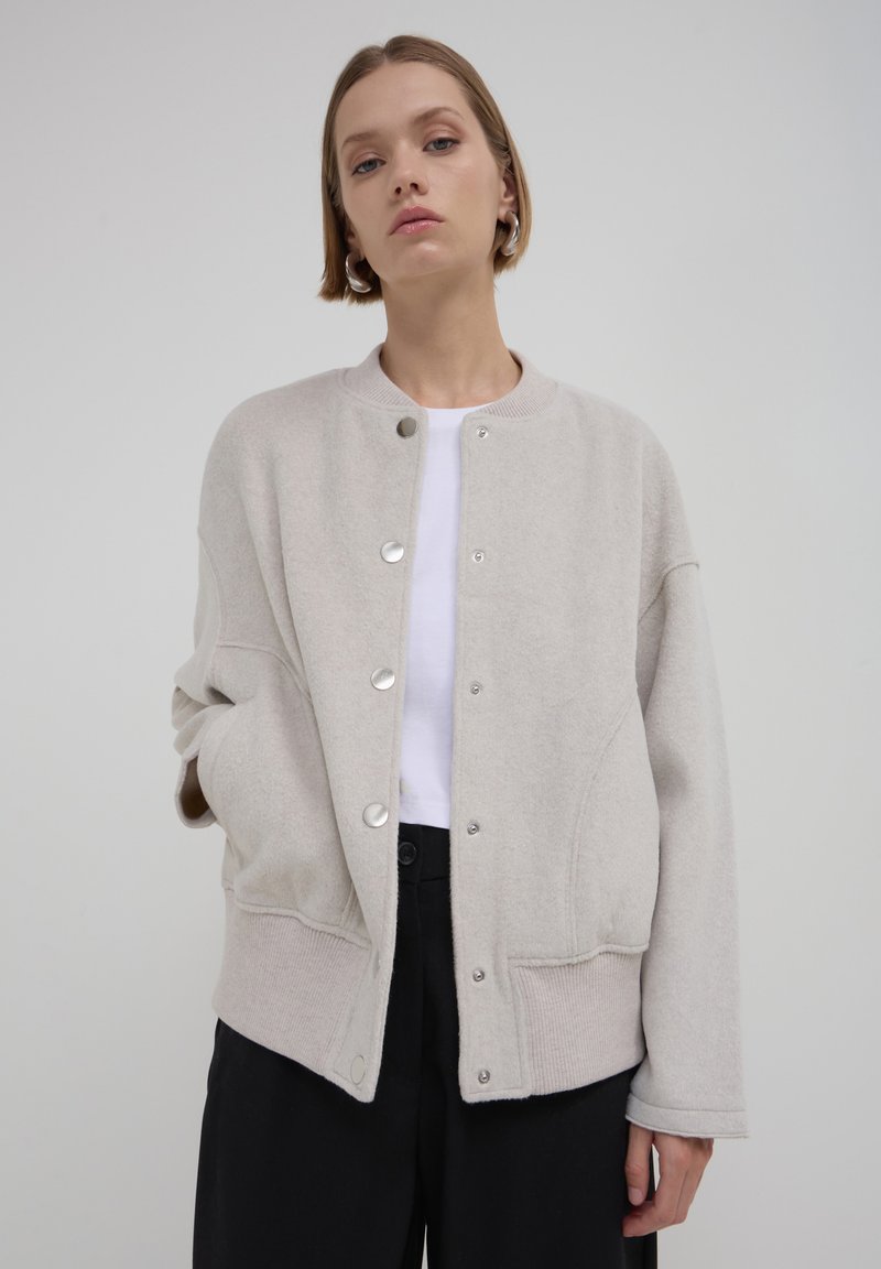 Veste en mélange de laine beige avec un ourlet côtelé, fermeture à pression et coutures courbes. Caractéristiques : coupe décontractée et épaules tombantes.