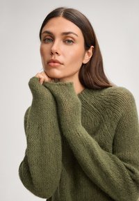 Maglione lavorato a maglia verde oliva con un motivo testurizzato, dotato di maniche lunghe e colletto alto. Materiale morbido e peloso con una vestibilità ampia.
