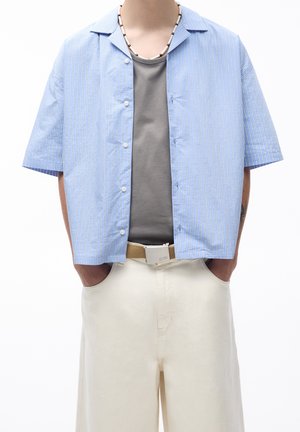 Chemise à manches courtes bleu clair avec des rayures verticales blanches, dotée d'un col et d'une fermeture à boutons, portée avec un t-shirt gris et un pantalon beige.