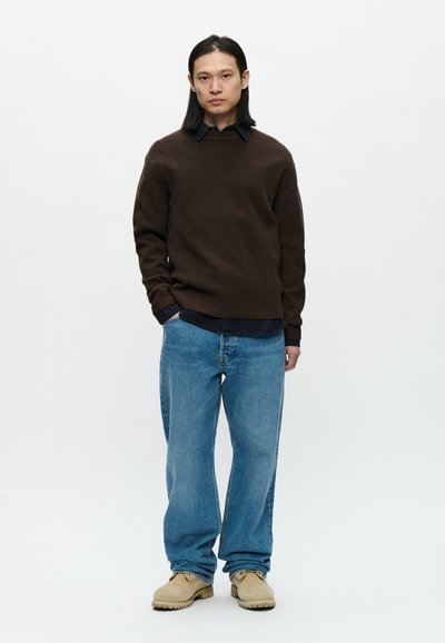 Suéter marrom texturizado sobre uma camisa azul escura, combinado com jeans claros e soltos e calçado bege. Design simples e casual.