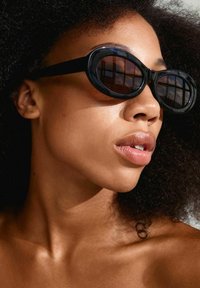 Black oval sunglasses with dark lenses, thick black frames. Model has curly hair and is positioned in soft lighting against a plain background.