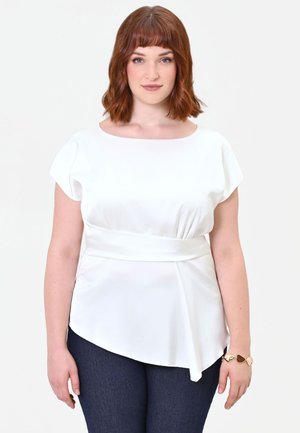 Donna con capelli rossi che indossa una camicetta bianca asimmetrica e pantaloni scuri, in piedi su uno sfondo chiaro e uniforme.