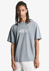 Grå bomulds t-shirt med korte ærmer og en afslappet pasform. Har hvid grafisk tekstdesign på fronten. Glat tekstur og rund halsudskæring.