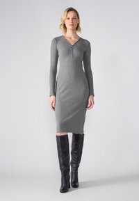 Robe en tricot côtelé gris avec un décolleté en V et des détails de fermeture éclair, associée à des bottes en cuir noir à hauteur de genoux. Texture lisse et design ajusté.
