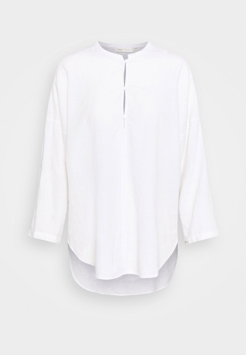 Camicia bianca in lino con scollatura rotonda, mezze bottoni parziali, maniche a tre quarti e orlo posteriore leggermente più lungo. Texture morbida.