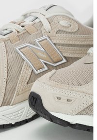 Beżowe buty sportowe z siateczkowymi i zamszowymi elementami. Posiadają wyraźne srebrne logo "N", teksturowane panele oraz czarną gumową podeszwę.