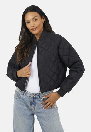 Femme portant une veste bomber matelassée noire sur un haut blanc et un jean bleu clair, posant avec une main sur la fermeture éclair et l'autre sur la hanche.
