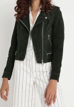 Kobieta w ciemnozielonej biker kurtce z zamszu z srebrnymi zamkami błyskawicznymi oraz białych spodniach w pionowe paski, stojąca na tle jednolitej ściany.