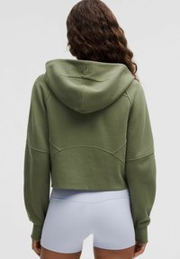 Hosszú göndör hajú nő olajzöld cropped kapucnis pulóverben és világosszürke testhezálló sportnadrágban áll, háttal a fényképezőgépnek.