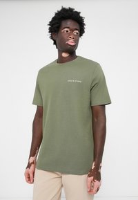 T-shirt à manches courtes vert olive en coton doux, avec un col rond et un petit logo blanc sur la poitrine. Design simple et épuré.