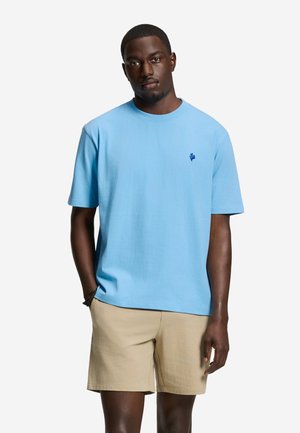 Man draagt een lichtblauw t-shirt met korte mouwen met een klein logo en beige korte broek, staat met één hand in zijn zak.