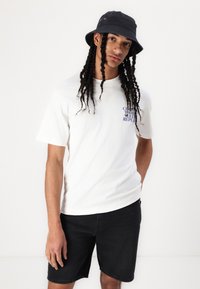 Weißes Baumwoll-T-Shirt mit blauer Druckgrafik; schwarze Denim-Shorts; schwarzer Bucket Hat. Das Model posiert mit den Händen in den Taschen vor einem neutralen Hintergrund.