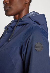 Giacca impermeabile blu navy con cappuccio con zip, cordini elastici e una toppa rotonda sul spalla sinistra. Tessuto texture.