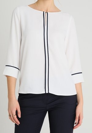 Weiße Bluse mit langen Ärmeln und schwarzem Besatz an den Manschetten und der Mittelnaht, kombiniert mit schwarzer Hose, abgebildet vom Hals bis zur Hüfte.