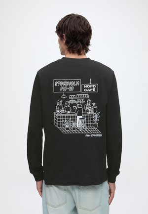 Hombre que lleva una camisa negra de manga larga que muestra una ilustración blanca de una escena de café con personas, plantas y carteles que dicen Stockholm Pop-Up y Mo’pd Café.