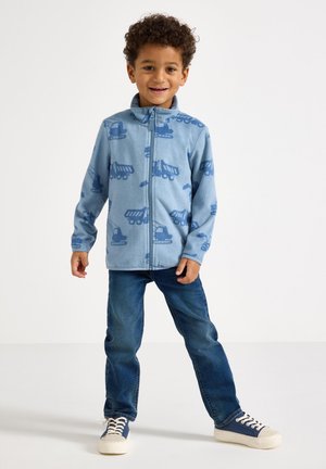 Giacca in pile azzurro chiaro con motivi di veicoli da costruzione blu, chiusura con zip e colletto alla coreana, abbinata a jeans di denim e sneakers blu.