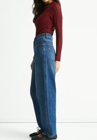 Red textured long-sleeve top paired with high-waisted, wide-leg blue jeans. Model wears black ballet flats. Simple, neutral background.
