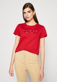 Raudona medvilninė marškinė su trumpomis rankovėmis, ant kurių yra sidabriniai "GUESS EST. 1981" logotipo puošimai. Derinama su smėlio spalvos pritaikytais kelnais.