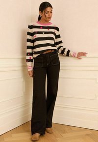 Pulover rayat în crem și negru cu accente roz și broderie; asociat cu blugi negri evazați cu talie înaltă și pantofi bej.
