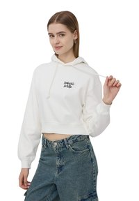 Hoodie bianco croppato con cappuccio con cordino e logo testuale ricamato in nero sul petto. Abbinato a jeans blu a vita alta.