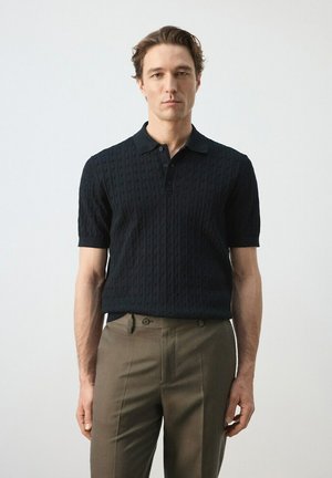 Joven de pie contra fondo blanco, vistiendo un polo de punto negro de manga corta y pantalones marrones a medida, mirando hacia adelante.