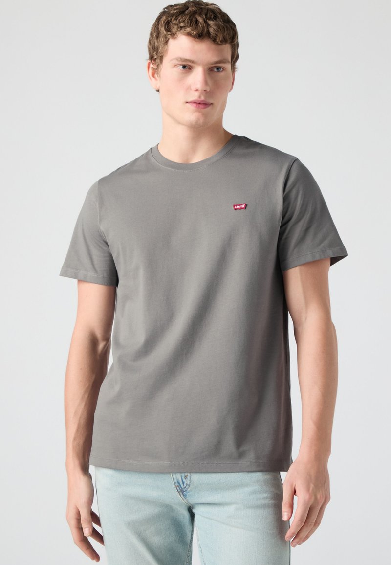 Jeune homme portant un t-shirt Levi's gris uni et un jean bleu clair, debout devant un fond clair uni.