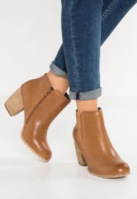 Stivaletti alla caviglia in pelle tan con chiusura a zip, punta rotonda e tacco impilato texturizzato. Indossati con jeans blu a risvolto.