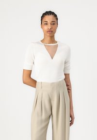 Bijela kratka majica s kratkim rukavima s V-izrezom i detaljima od prozirnog materijala. Uparena s bež, plisiranim hlačama visokog struka. Glatka tekstura, prilagođen dizajn.