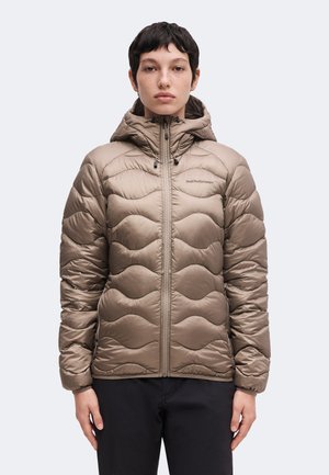 Persoon met kort donker haar draagt een taupekleurige gewatteerde jas met capuchon en een donkere broek, staand frontaal op een effen achtergrond.