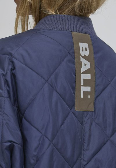Marineblå quiltet jakke med ribbestrikket krage. Har en vertikal brun lapp med ordet "BALL" i hvitt. Jevn, reflekterende tekstur.